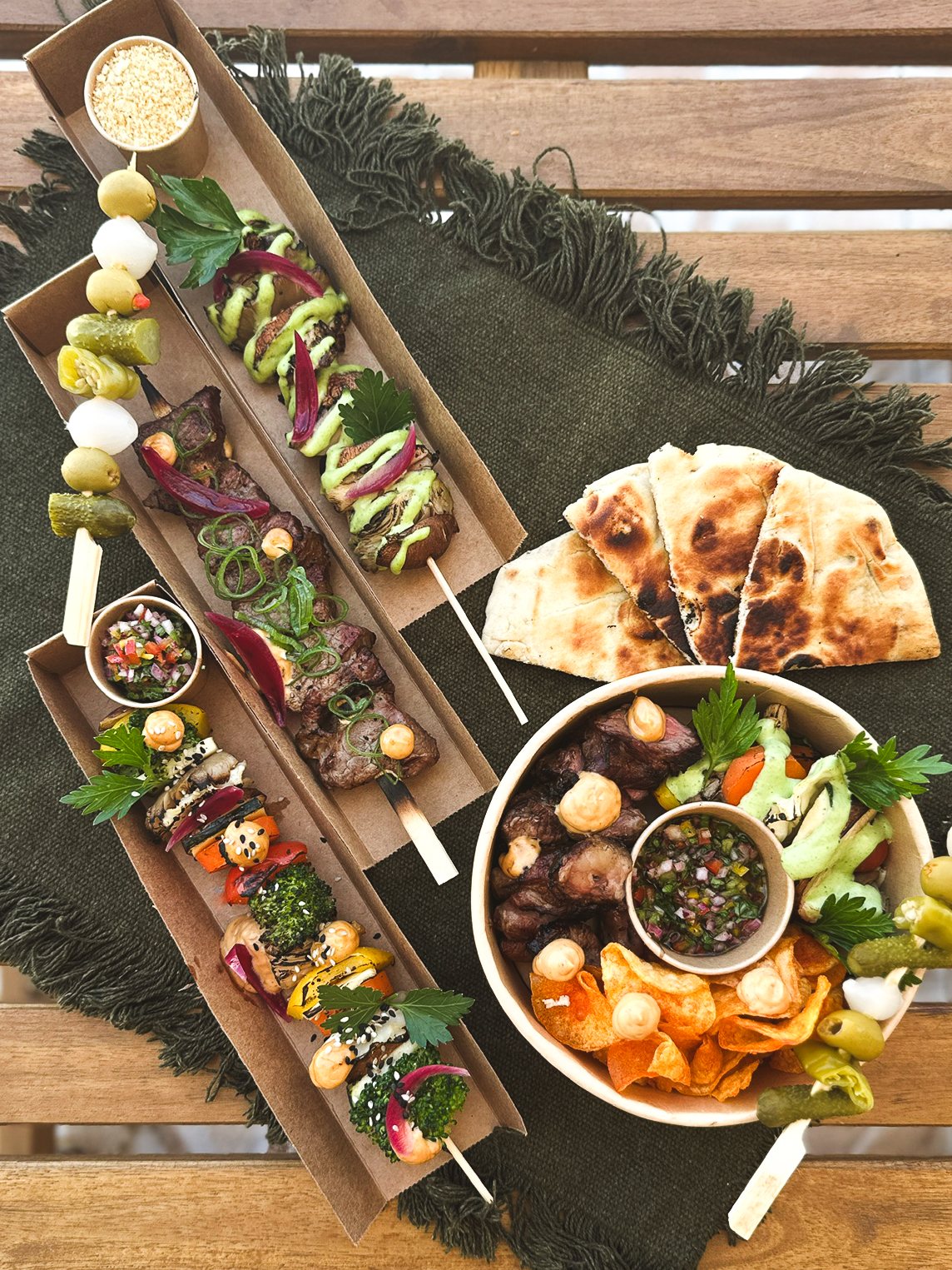 Caixas de street food com espetos de carne e vegetais, pão grelhado e um bowl com chips